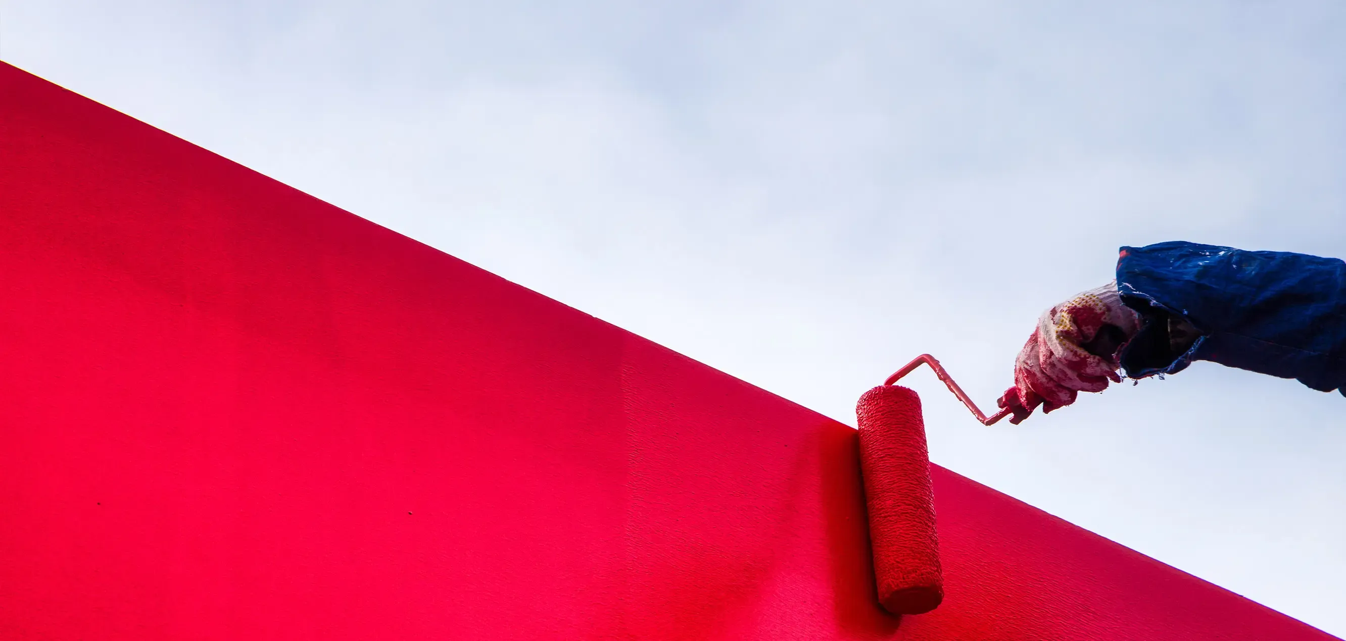 Eine Person in blauer Kleidung trägt einen Farbroller mit roter Farbe. Der Farbroller berührt eine rot gestrichene Wand, die sich schräg nach oben erstreckt. Der Himmel im Hintergrund ist hell und bewölkt.