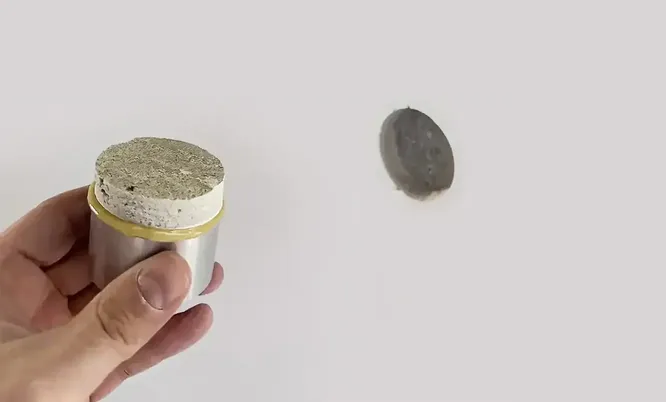 Hand hält eine bei der Haftzugprüfung entnommene runde Materialprobe vor der entsprechenden Prüf- und Entnahmestelle an einer Wand.
