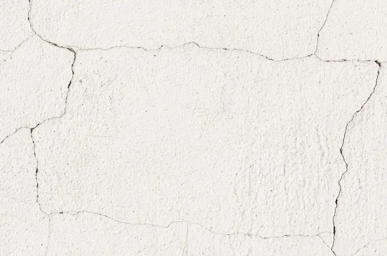 Eine Nahaufnahme einer weißen Wand mit mehreren Rissen und einer rauen Textur. Die Risse verlaufen unregelmäßig und erzeugen ein erodiertes Aussehen.
