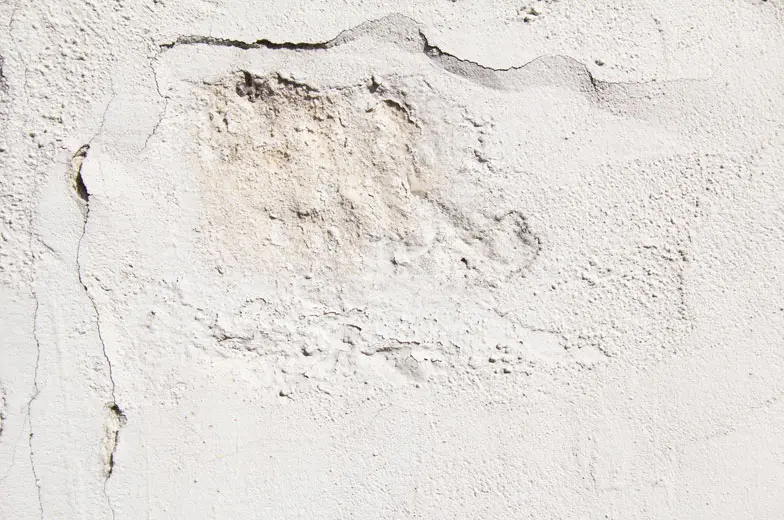 Unebene, strukturierte weiße Wand mit sichtbaren Rissen und Stellen unterschiedlicher Textur. Einige Bereiche sind dunkler oder rauer, was der Oberfläche ein gealtertes Aussehen verleiht.