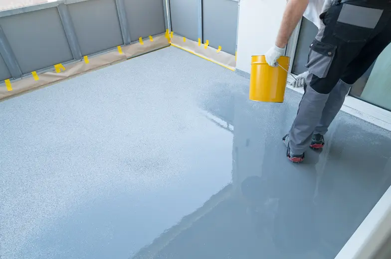 Eine Person trägt mit einem gelben Eimer eine graue Flüssigkeit auf einen Boden auf, wahrscheinlich zur Versiegelung. Der Boden ist zur Hälfte bearbeitet. Schutzfolie und gelbes Klebeband sind am Rand angebracht.
