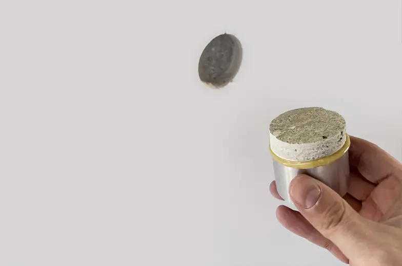 Eine Hand hält einen Metallzylinder mit offenem Ende und einer runden Öffnung, aus der eine Probe sichtbar ist. Im Hintergrund befindet sich eine runde Öffnung oder Vertiefung in einer Wand.