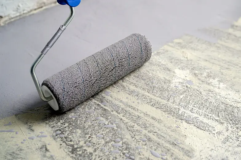 Ein grauer Farbroller wird über eine strukturierte Oberfläche gerollt, um sie mit grauer Farbe zu beschichten. Der Untergrund zeigt vorherige unregelmäßige Farbschichten und der Farbroller hat einen blauen Griff.