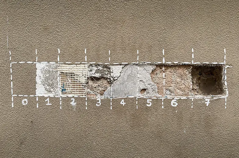 Eine Wand mit rechteckigem Ausschnitt, der verschiedene Schichten sichtbar macht, darunter Putz, Netze und Mauerwerk. Die Bereiche sind mit nummerierten Markierungen von 0 bis 7 umrandet.