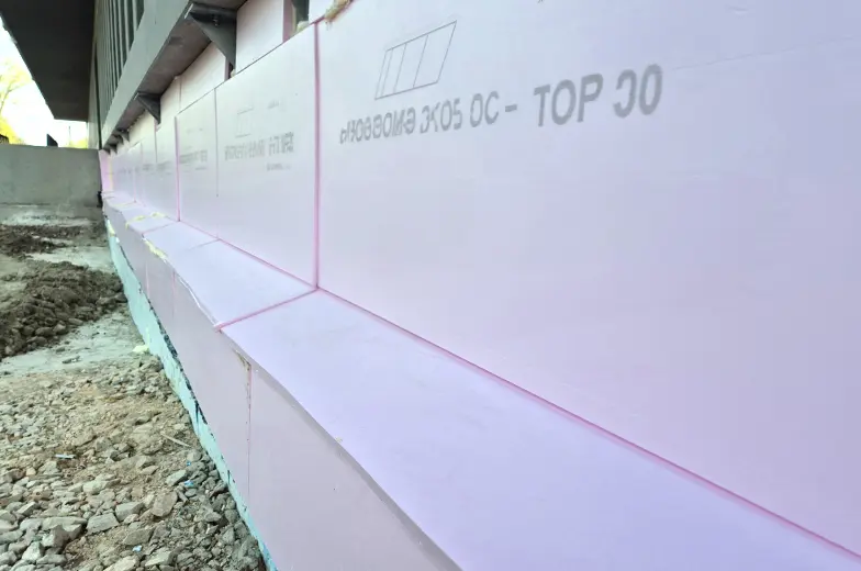 Ein Gebäude wird mit rosa Dämmplatten isoliert. Die Platten sind auf der Außenwand angebracht. Der Boden ist mit Erde bedeckt und der Fokus liegt auf der Isolierung.