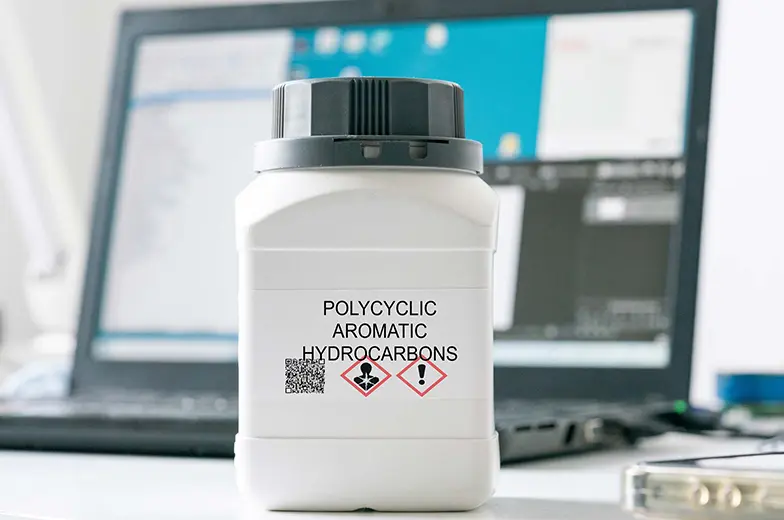 Ein weißer Behälter mit der Aufschrift "POLYCYCLIC AROMATIC HYDROCARBONS" steht im Vordergrund. Zwei Gefahrensymbole sind darauf abgebildet. Im Hintergrund unscharf ein Laptop und Bildschirm.