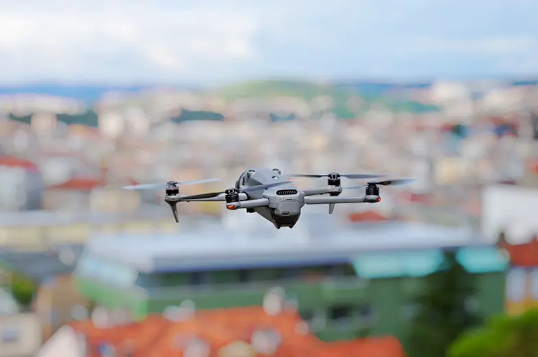 Eine Drohne fliegt über einer unscharfen Stadtlandschaft. Der Himmel ist teilweise bewölkt. Die Drohne hat vier Propeller und eine Kamera an der Vorderseite. Im Hintergrund sind Gebäude in verschiedenen Farben.