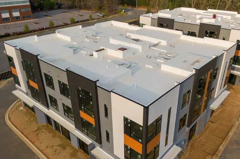 Ein modernes, großes Gebäude mit Flachdach und mehrfarbiger Fassade steht auf einer Baustelle. Es hat große, getönte Fenster, vertikale schwarze Akzente und orangefarbene Streifen an den Ecken. Im Hintergrund sind Bäume und Parkplätze sichtbar.