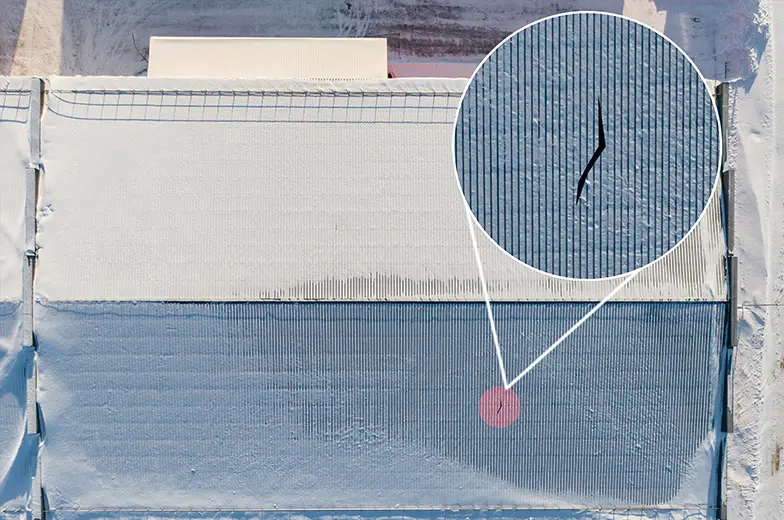 Luftaufnahme einer schneebedeckten Fläche mit parallelen Linien. Ein vergrößerter Kreis zeigt einen kleinen Riss im Schnee. Oben ist eine Struktur, möglicherweise ein Gebäude, teilweise sichtbar.