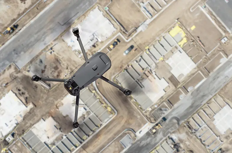 Eine Drohne fliegt über einer Baustelle mit mehreren umrandeten Bereichen und Gebäuden. Der Blick ist von oben, und die Drohne ist das zentrale Element im Bild. Man sieht Straßen und Bauflächen.